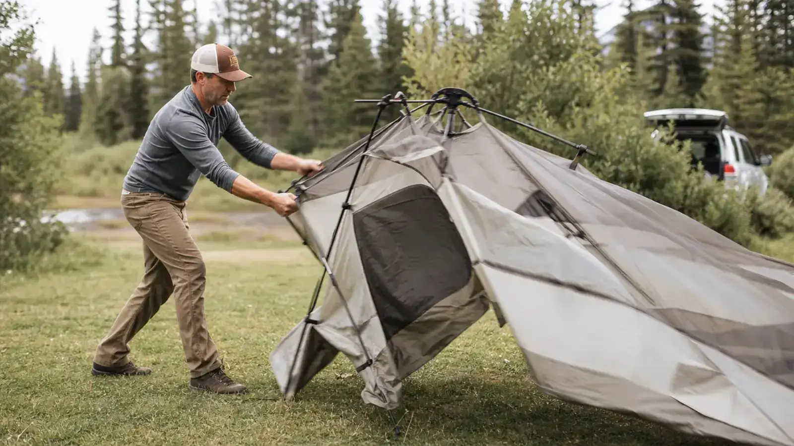 A man unfolding a large green family camping tent at a forested campsite on a sunny afternoon, poles spreading outward mid-setup