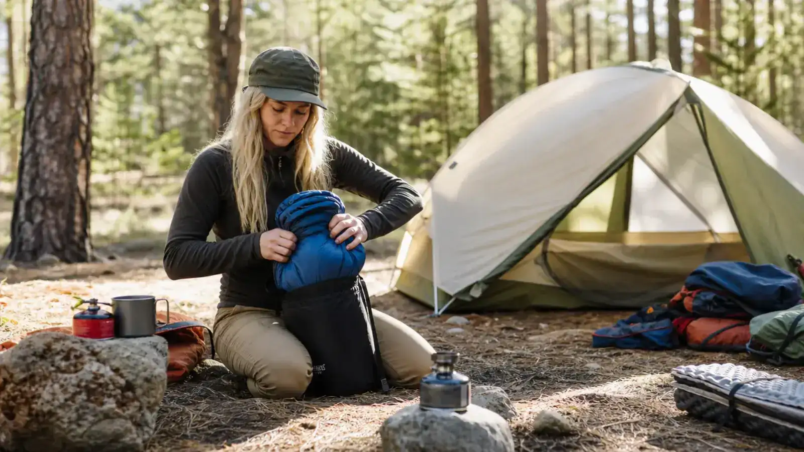 Camper at a forested campsite packing gear into a car trunk with sleeping bag rolled up nearby